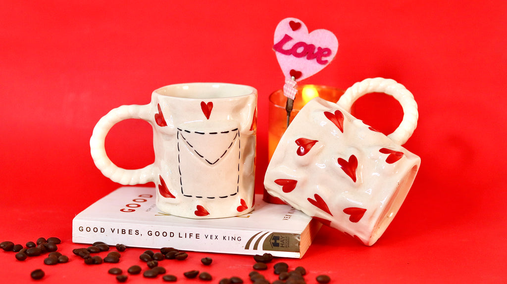Two ceramic mugs with heart designs on a red background, accompanied by coffee beans and a 'Love' candle.