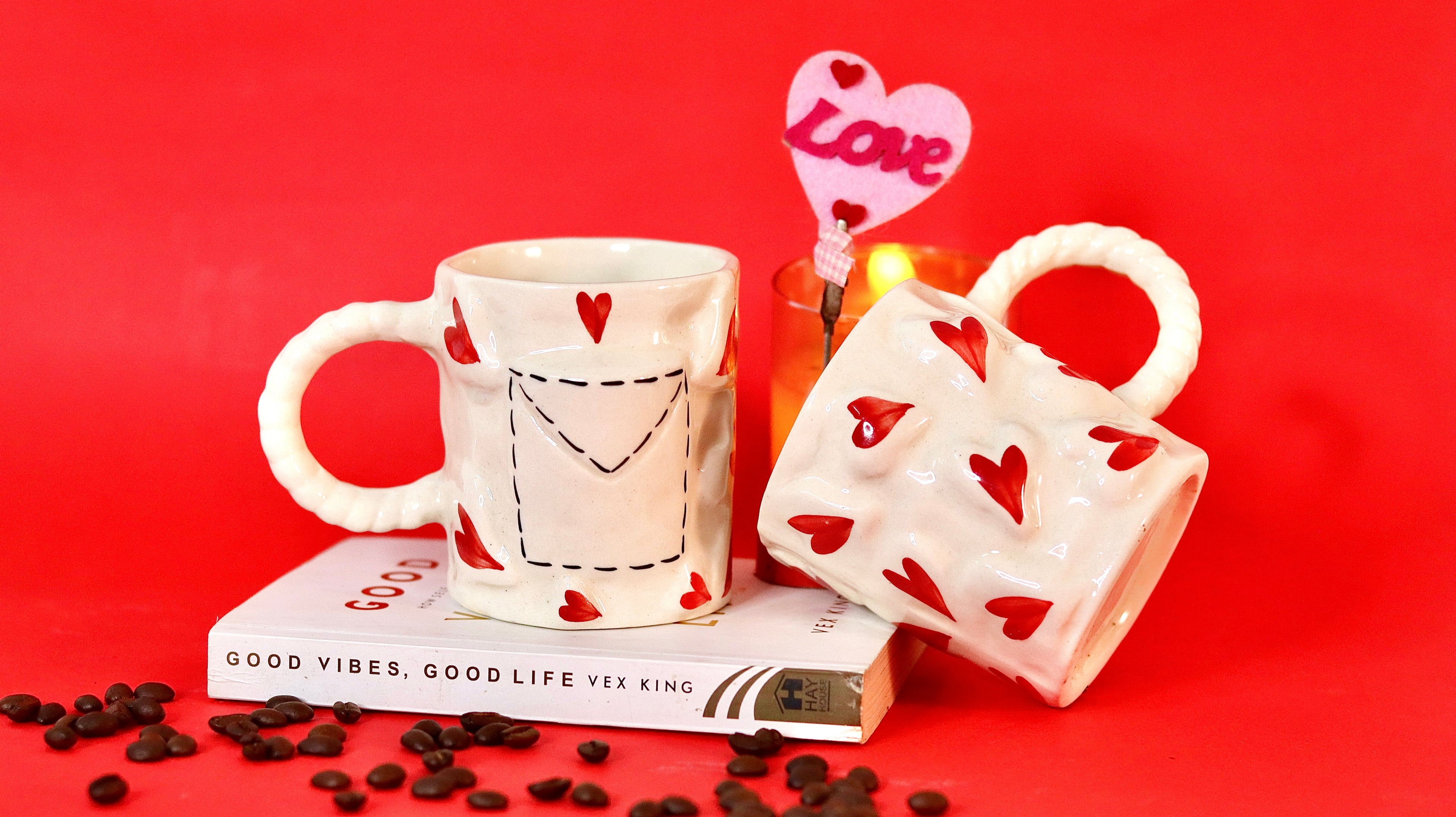 Two ceramic mugs with heart designs on a red background, accompanied by coffee beans and a 'Love' candle.