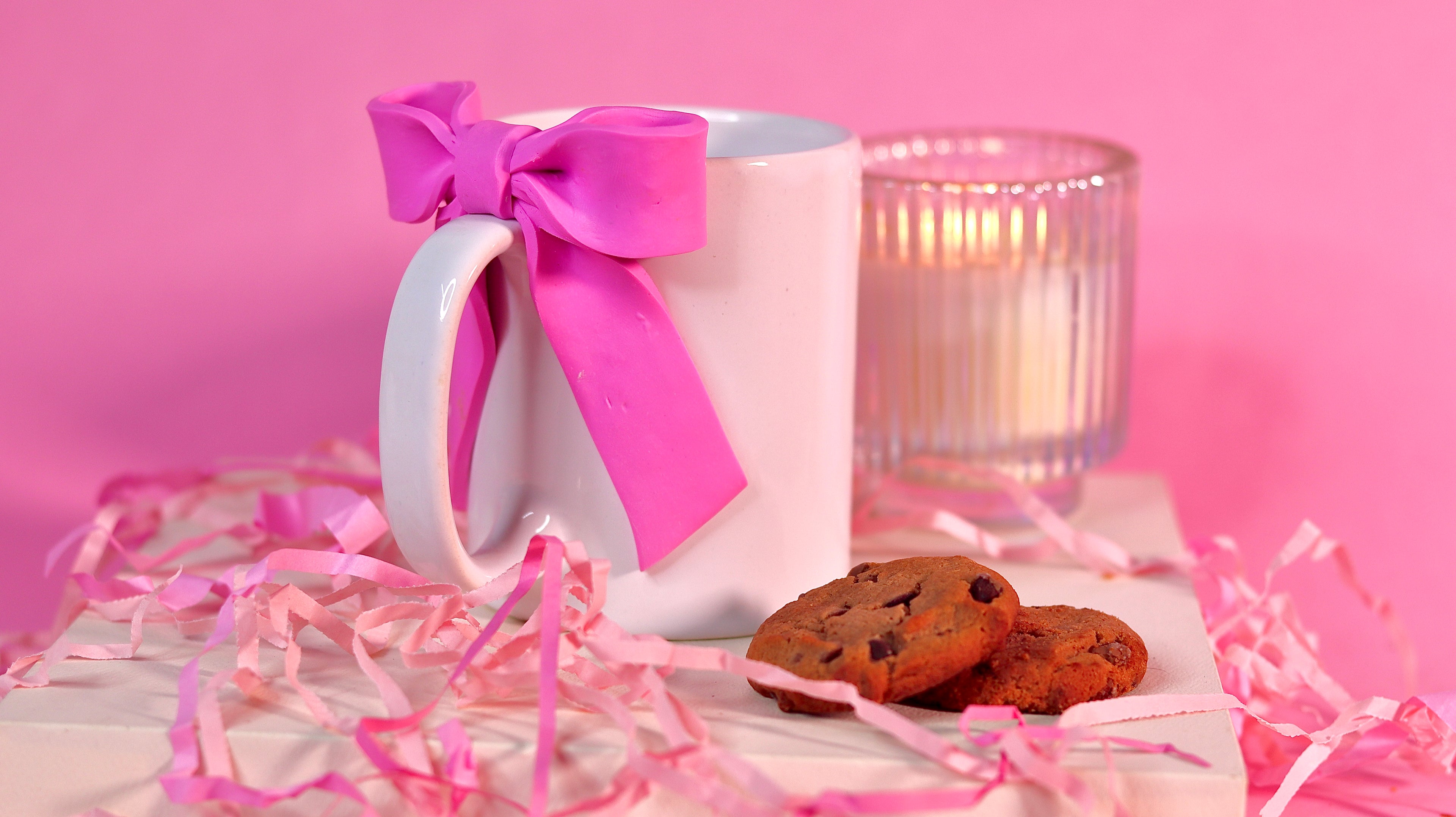 White mug with pink bow, cookies, and candle on a pink background